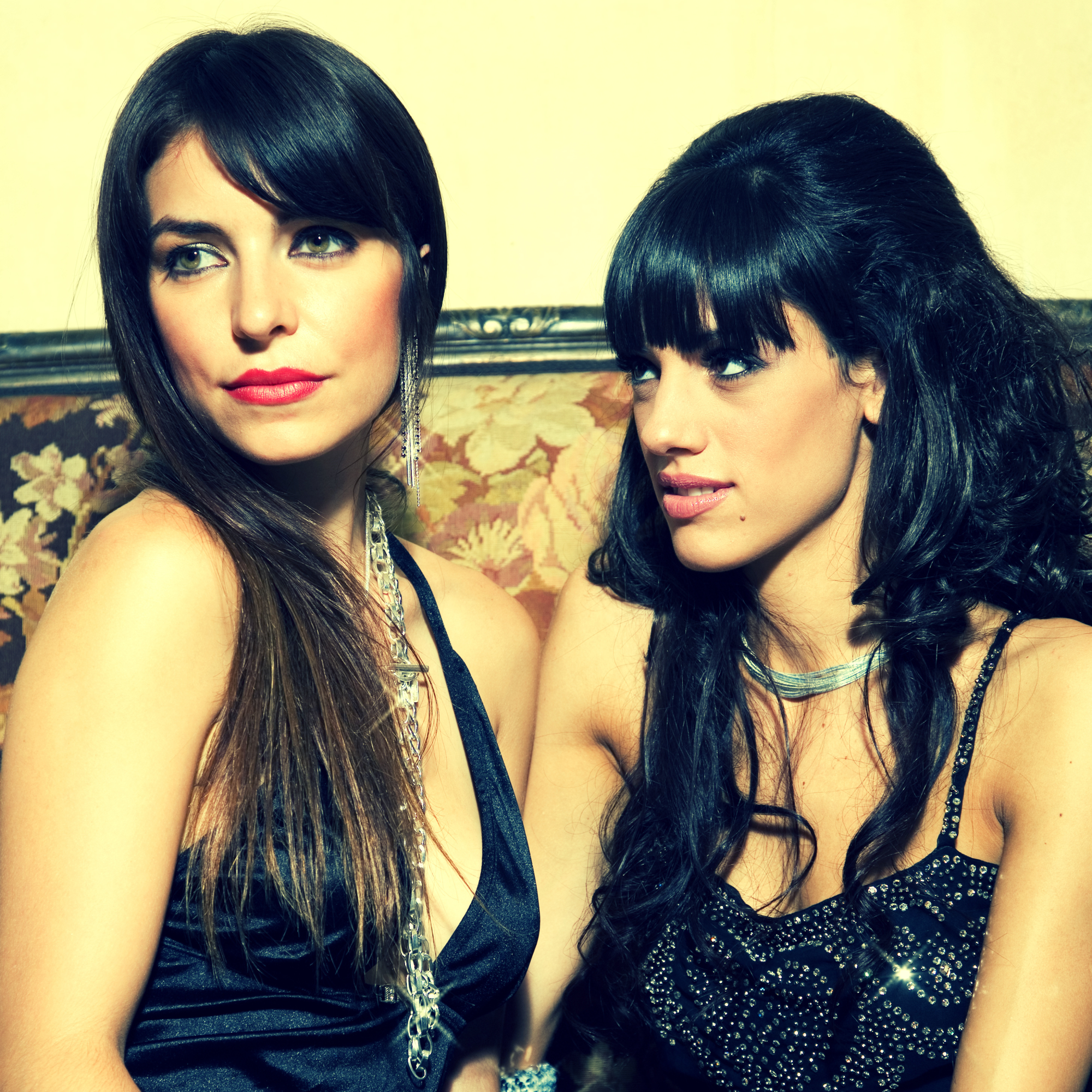 two women sitting on a couch wearing black cocktail attire, presumably looking at people around them at a social event